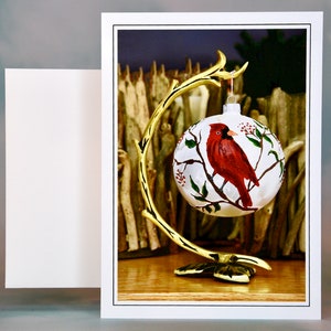 Puede incluir: Una bola de Navidad blanca con un diseño de cardenal rojo posado en una rama con hojas verdes y bayas rojas. La bola cuelga de un soporte de metal dorado. A la izquierda hay una tarjeta blanca en blanco.