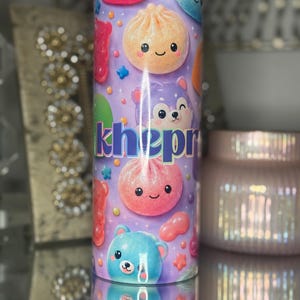 May include: A cylindrical tumbler with a purple background featuring a pattern of colourful Nee Doh stress balls, including round and bear-shaped designs with smiling faces. The name "khepr" is printed in bold, dark blue letters across the centre.