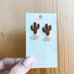 Op de afbeelding: Een paar bruine cactus oorbellen met gouden zonnestralen accenten. De oorbellen worden getoond op een witte kaart met de tekst "lively honey" en een logo van een cactus en een zon.