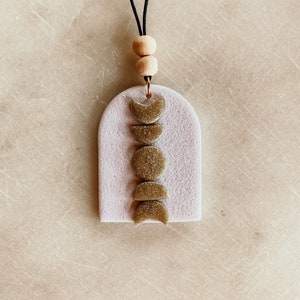 May include: A white ceramic car air freshener with a gold glitter moon phase design. The air freshener is attached to a black cord with two wooden beads.