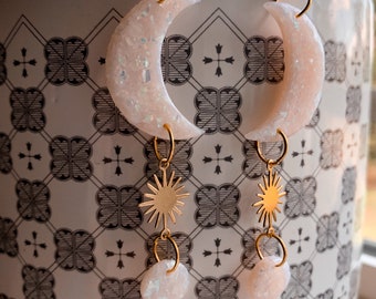 Puede incluir: Un par de pendientes de oro con un encanto de media luna y estrella. La luna está hecha de resina blanca con purpurina y la estrella es de metal dorado.