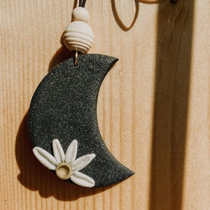 以下が含まれることがあります： 白いデイジーの花がついた黒い三日月形のチャームが、白いビーズのついた茶色の紐にぶら下がっています。