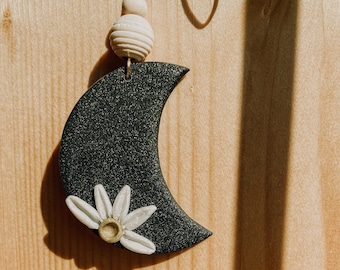 May include: A black crescent moon charm with a white daisy flower, hanging from a brown cord with white beads.