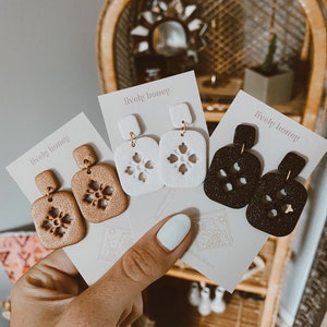 Op de afbeelding: Drie paar oorbellen met een sneeuwvlokdesign. De oorbellen zijn gemaakt van polymer clay en verkrijgbaar in bruin, wit en zwart. De oorbellen worden gepresenteerd op witte kaartjes met de tekst "lively honey" erop gedrukt.