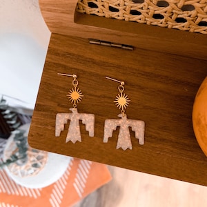 May include: A pair of gold-colored earrings featuring a sunburst design at the top and a white, bird-shaped pendant with glitter accents. The earrings are displayed on a dark wooden surface.