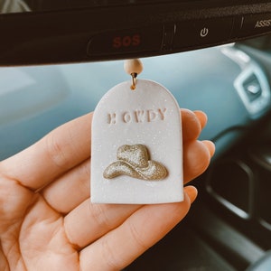 May include: A white, glittery car air freshener with a rounded top and a gold cowboy hat design. The word "HOWDY" is embossed in pink letters above the hat. A wooden bead and string are attached for hanging.