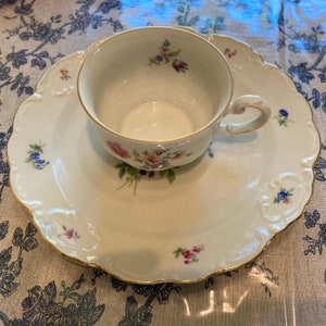 May include: A white porcelain teacup and saucer set with a floral pattern. The cup has a gold rim and the saucer has a scalloped edge.