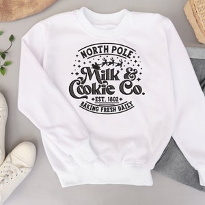May include: White sweatshirt with a black graphic design that says "North Pole Milk & Cookie Co. Est. 1802 Baking Fresh Daily" with a Santa and reindeer illustration.