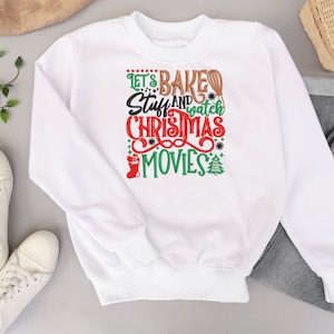Pu&ograve; includere: Felpa bianca con un design natalizio festoso che dice "Let's Bake Stuff and Watch Christmas Movies". Il design presenta una calza rossa, un albero di Natale verde e una frusta marrone.