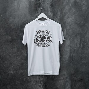 May include: White t-shirt with a black graphic design that says "North Pole Milk & Cookie Co. Est. 1802 Baking Fresh Daily".