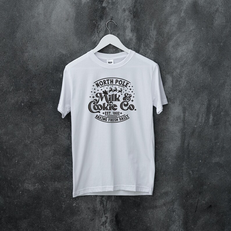 May include: White t-shirt with a black graphic design that says "North Pole Milk & Cookie Co. Est. 1802 Baking Fresh Daily".