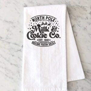 May include: White kitchen towel with a black and white design that says "North Pole Milk & Cookie Co. Est. 1802 Baking Fresh Daily". The design features a Santa Claus in a sleigh with reindeer flying through the air.