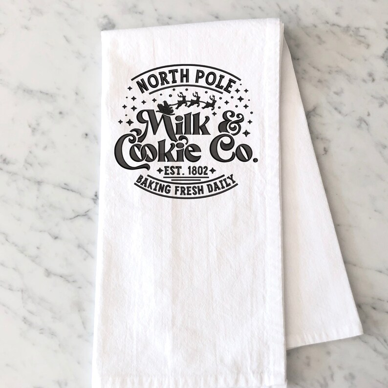 May include: White kitchen towel with a black and white design that says "North Pole Milk & Cookie Co. Est. 1802 Baking Fresh Daily". The design features a Santa Claus in a sleigh with reindeer flying through the air.