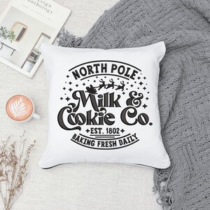 May include: White throw pillow with black embroidery that reads "North Pole Milk & Cookie Co. Est. 1802 Baking Fresh Daily". The design includes a silhouette of Santa Claus in a sleigh with reindeer.