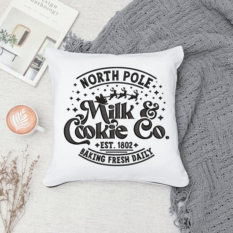 May include: White throw pillow with black embroidery that reads "North Pole Milk & Cookie Co. Est. 1802 Baking Fresh Daily". The design includes a silhouette of Santa Claus in a sleigh with reindeer.
