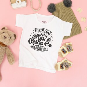 May include: White baby onesie with a black and white graphic that says "North Pole Milk & Cookie Co. Est. 1802 Baking Fresh Daily".