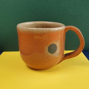 Op de afbeelding: Een gespikkelde oranje keramische mok met een groen en bruin cirkelontwerp. De mok heeft een afgerond lichaam en een handvat.