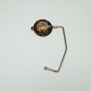 May include: A silver-toned handbag hook with a round, brown turtle design. The turtle has a yellow and orange shell.