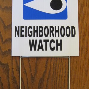 May include: A white sign with the words "WARNING NEIGHBORHOOD WATCH" in black text. The sign has a blue rectangle with a white eye symbol. The sign is mounted on a metal stake.