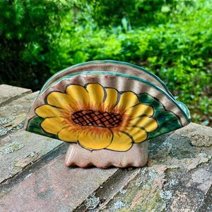 May include: Ceramic napkin holder shaped like a sunflower with yellow petals, a brown center, and green leaves. The holder is painted in a rustic style and has a light brown base.