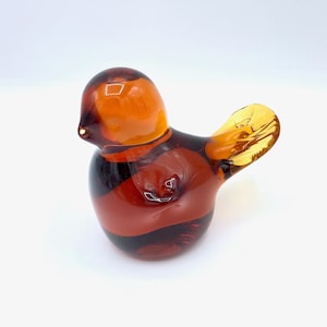 May include: A small amber glass bird figurine with a rounded body and a long, thin tail. The bird is a light amber color with darker amber accents.