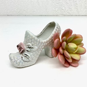 May include: A white ceramic shoe planter with a pink rose detail. The shoe has a high heel and is decorated with a textured surface. A pink and green succulent is placed next to the shoe.