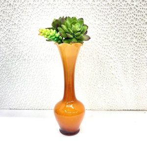 May include: An amber-colored glass vase with a flared, scalloped rim. The vase holds a cluster of green and yellow artificial succulents. The vase has a bulbous base and a slender neck, standing approximately 10 inches tall. The background is a textured white surface.