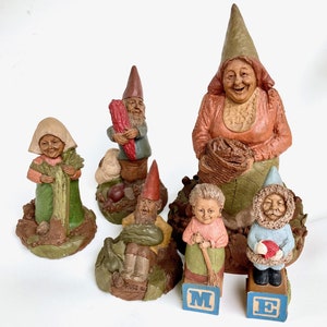 May include: A collection of whimsical garden gnome figurines. The gnomes are depicted in various poses, holding vegetables, baskets, and brooms. They wear traditional pointed hats and colorful clothing. The figurines are made of a textured material with detailed facial features.