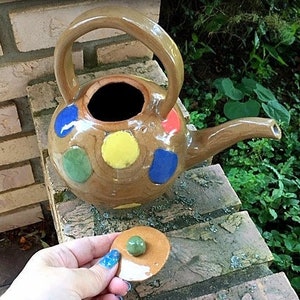 May include: A brown ceramic teapot with a colorful polka dot design. The teapot has a lid and a spout. The teapot is sitting on a brick surface.