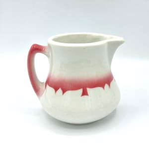 May include: A small, white ceramic pitcher with a red handle and a red band around the middle. The red band has a scalloped edge. The pitcher has a spout for pouring.