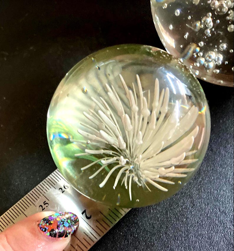 May include: Clear glass paperweight with a white floral design encased within. The paperweight is round and measures approximately 2 inches in diameter. Bubbles are visible within the glass, adding to its decorative appeal.