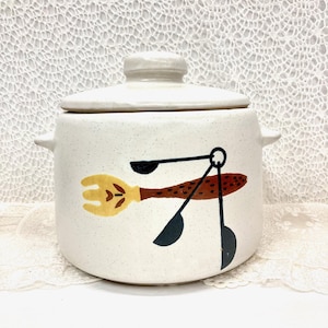 May include: White ceramic crock with a lid, featuring a brown, yellow, and black design of a fork, spoon, and measuring spoons.