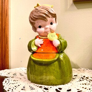 May include: A vintage ceramic cookie jar in the shape of a child holding a lollipop. The jar features a green dress, orange top, and brown hair. The text "Lollipops" is visible on the jar.