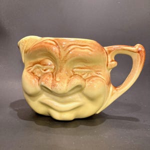 May include: A vintage ceramic pitcher with a smiling face design. The pitcher features a light yellow base color with brown accents on the face and handle. The face has closed eyes and a wide, smiling mouth. The handle is curved and attached to the side.