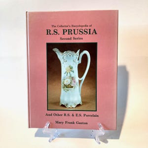 May include: A pink book titled "The Collector's Encyclopedia of R.S. PRUSSIA Second Series" with a picture of a white pitcher with floral designs on the front cover.