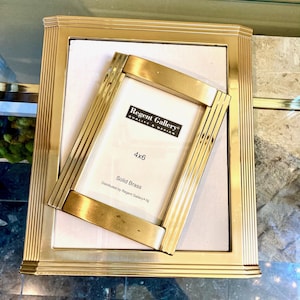 May include: A rectangular solid brass picture frame with a ribbed border design, featuring a smaller, angled solid brass frame on top. The inner white insert displays the text Regent Gallery, 4x6, and Solid Brass in black lettering.