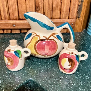 May include: A vintage ceramic pitcher and two matching salt and pepper shakers with a fruit design. The pitcher has a blue and white rim and features a red apple, yellow pear, and green leaves. The salt and pepper shakers are smaller versions of the pitcher with the same design.
