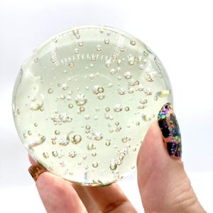 May include: A clear, spherical glass paperweight filled with numerous air bubbles. The paperweight has a light yellow tint and is held in a hand with a decorated nail. The bubbles vary in size, creating a unique visual effect.