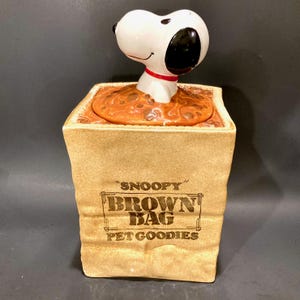 May include: A ceramic Snoopy cookie jar shaped like a brown paper bag. The bag reads "Snoopy Brown Bag Pet Goodies." Snoopy's head and neck emerge from the top, with a red collar. The jar has a brown lid with a textured surface.