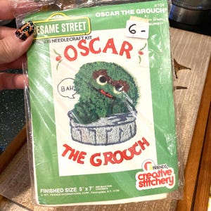 May include: A vintage Sesame Street Friends needlecraft kit featuring Oscar the Grouch. The kit includes a design of Oscar on a trash can, with the text "OSCAR THE GROUCH". Finished size is 5" x 7".