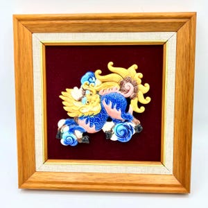 May include: A framed, colorful ceramic sculpture of a mythical creature. The sculpture features yellow, blue, and pink hues, set against a deep red background. The frame is made of light brown wood with a cream-colored inner border.