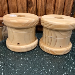 May include: Two cylindrical light-colored wooden spools with a smooth finish and visible grain patterns. Each spool features a wide top and bottom rim, a central circular opening, and a small hole near the base. A paper tag is attached to one side.