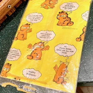 May include: Yellow Garfield-themed wrapping paper with multiple images of Garfield in various poses. Speech bubbles contain phrases like "Happy Birthday" and "Here's your gift." The paper is sealed in a clear plastic wrapper.