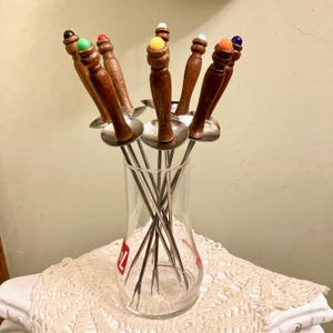 May include: A set of vintage fondue forks with wooden handles and colorful tips, displayed in a clear glass vase. The forks have stainless steel tines and small metal guards. The vase sits on a white lace tablecloth.