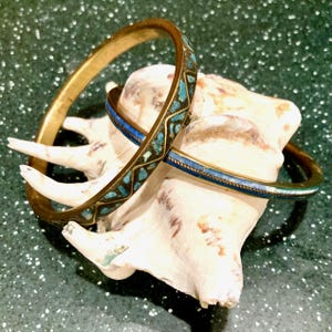 May include: Three brass bangles with turquoise and blue inlay rest on a white seashell. One bangle has a geometric turquoise pattern, while the others feature blue and white stripes. The bangles are displayed on a speckled green surface.