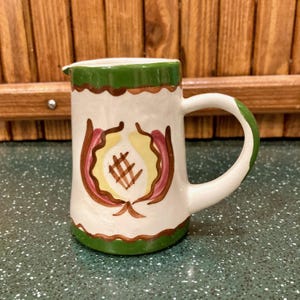 May include: A white ceramic pitcher with a green rim, base, and handle accents. The pitcher features a central design with yellow, brown, and pink floral elements. The handle is curved, and the pitcher has a small spout.
