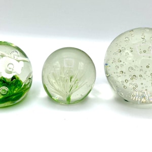 May include: Three clear glass paperweights. One features white flowers and green leaves, another has a white plant design, and the third is filled with bubbles. Decorative spheres for home decor.
