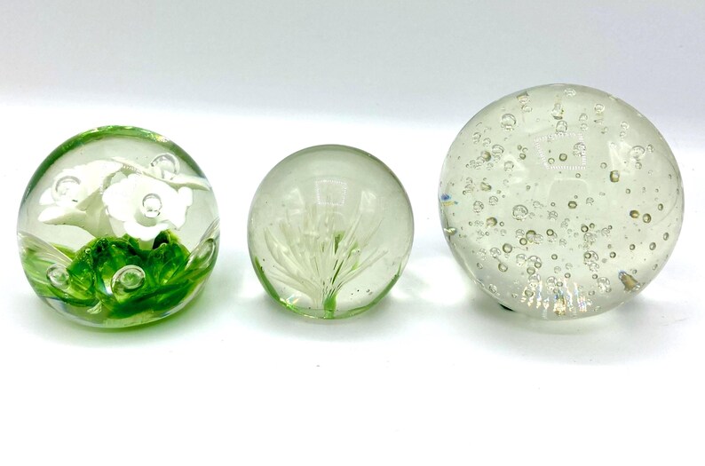 May include: Three clear glass paperweights. One features white flowers and green leaves, another has a white plant design, and the third is filled with bubbles. Decorative spheres for home decor.