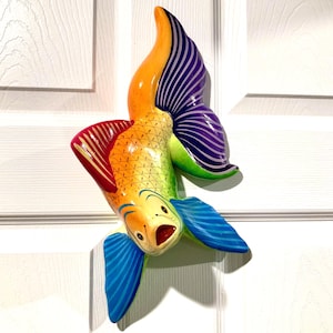 May include: A colorful, hand-painted fish sculpture with a whimsical design. The fish features a vibrant palette of orange, yellow, green, red, blue, and purple. The sculpture is likely made of wood or a similar material and is designed to be hung on a wall.