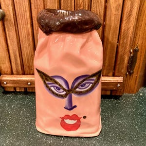 May include: A pink ceramic vase with a face design. The face features a purple mask, eyes, nose, and red lips. The top of the vase is brown. The vase is a unique decorative item.